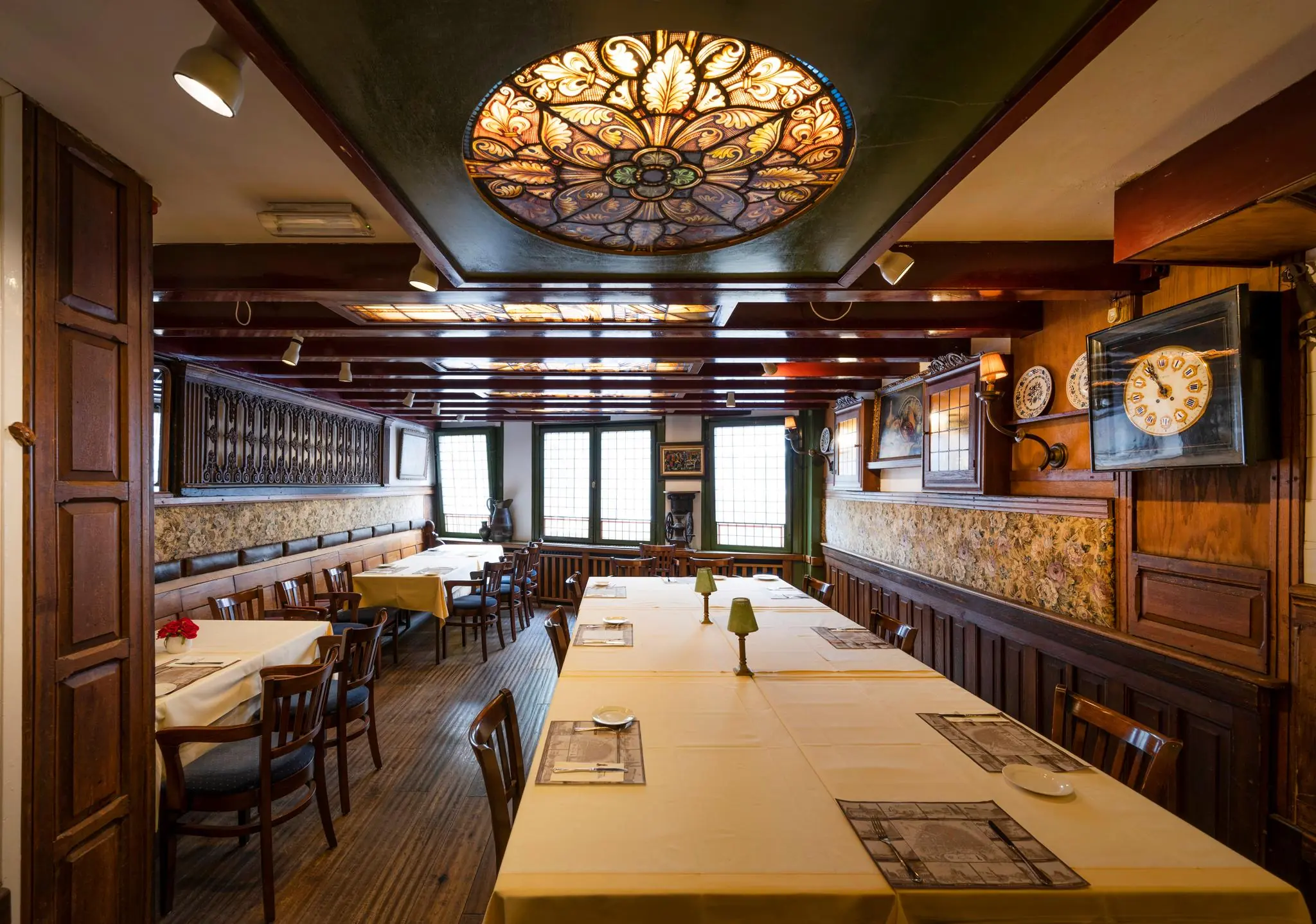 Gezellig ingericht restaurant aan de Nieuwezijds Voorburgwal met houten lambrisering, gedekte tafels en een kleurrijk glas-in-lood plafond.